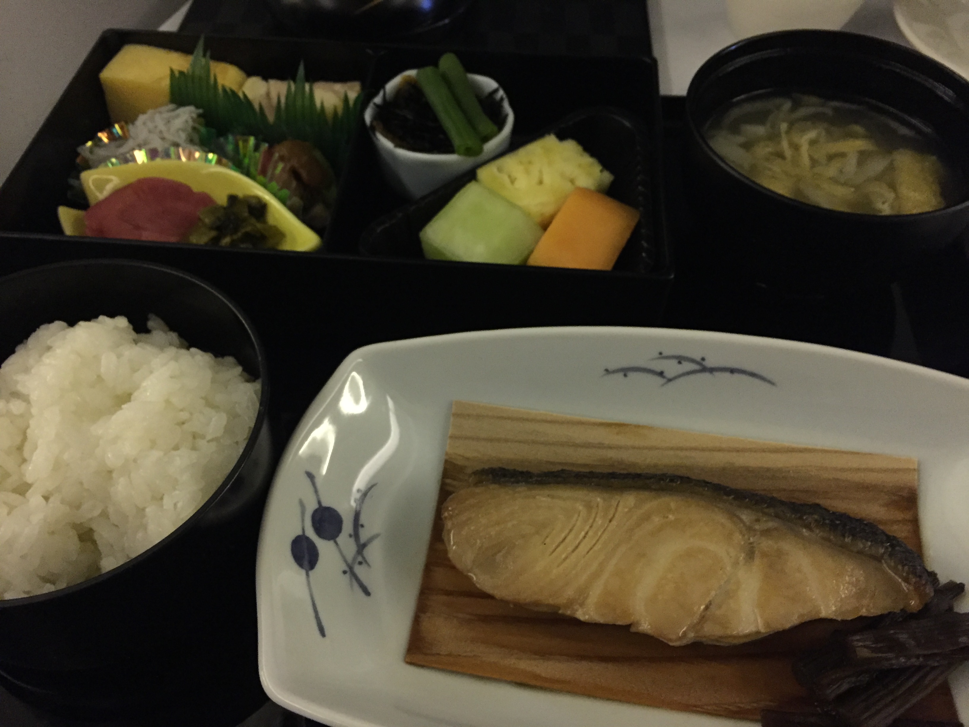 Japanese breakfast tray — grilled mackerel on a wood plank, rice, miso soup, tamagoyaki, pickles, and fruit