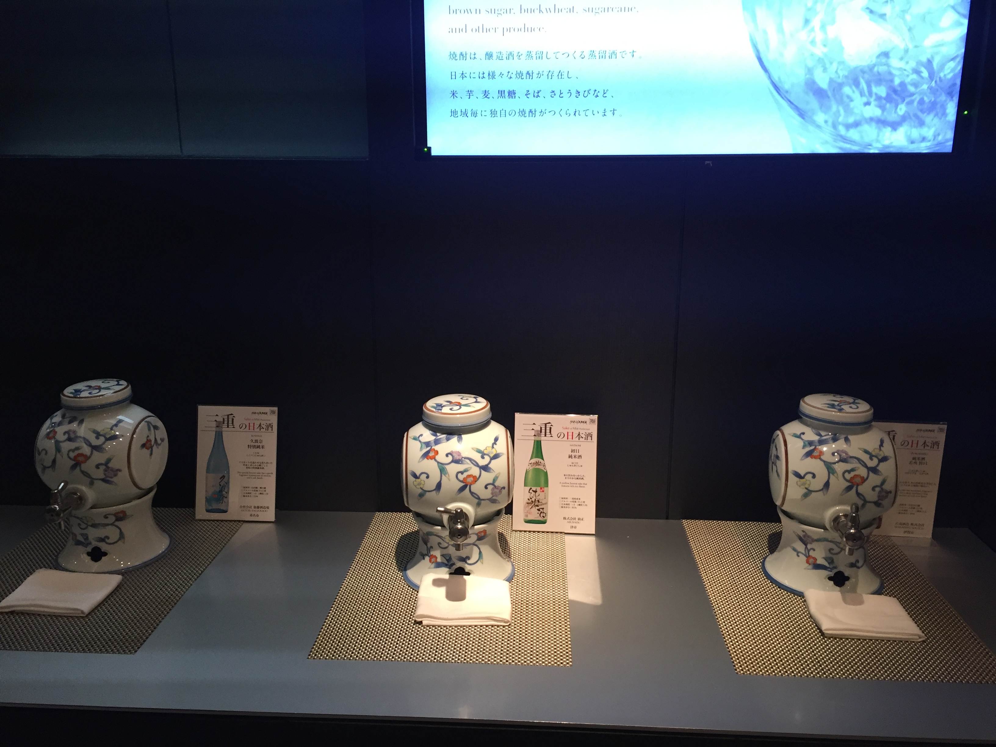 A sake exhibition — three porcelain dispensers on a black backdrop with information cards and a video presentation about brewing