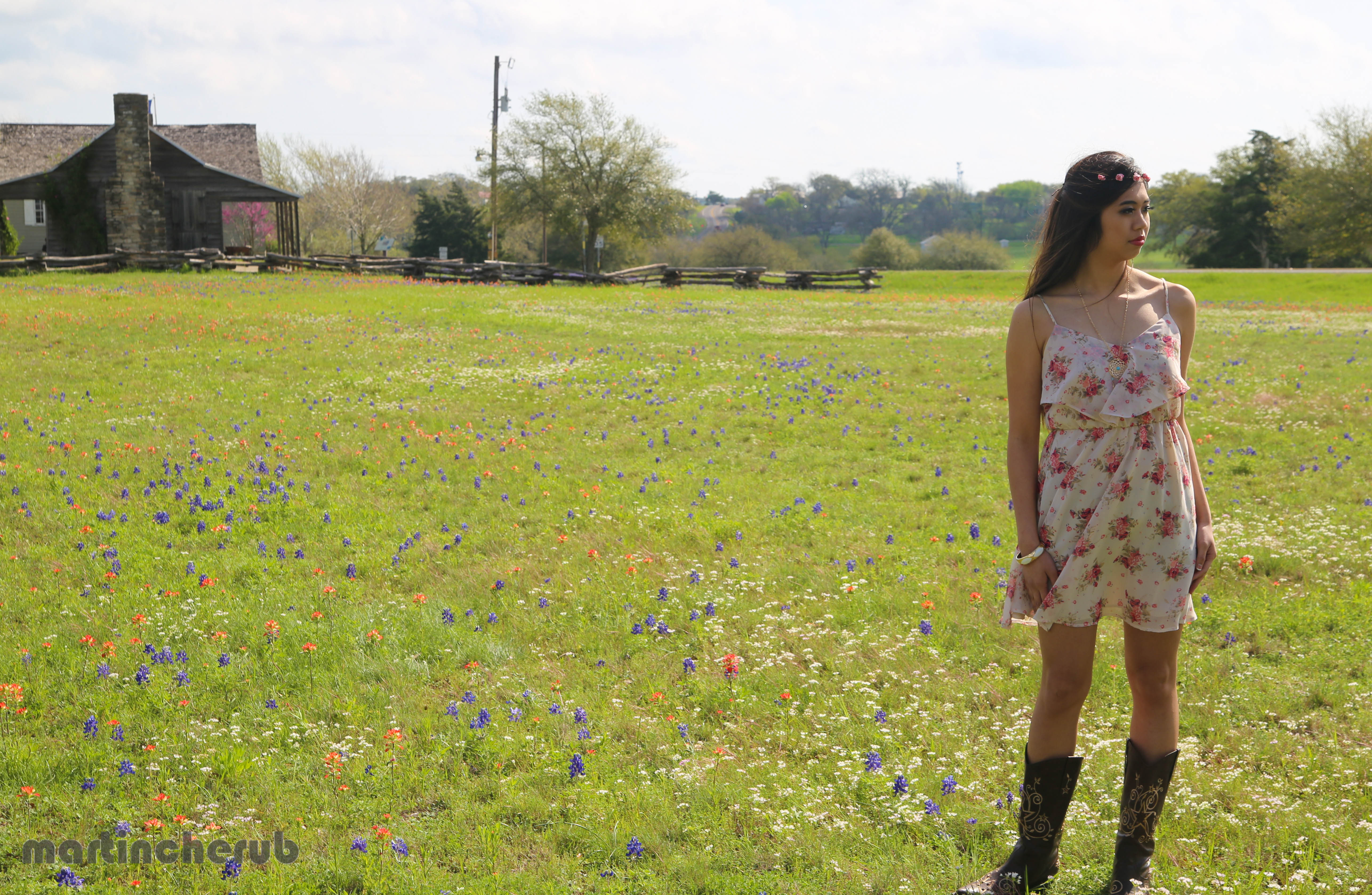 Tiffany Dimaano in a floral dress and cowboy boots, standing in the wildflower field with a historic log cabin in the background