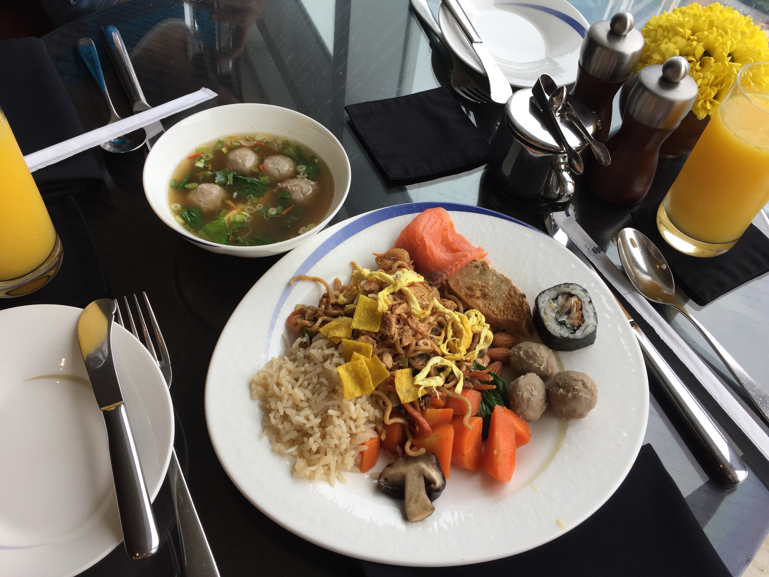 A breakfast plate of fried rice, mee goreng, sushi roll, mushrooms, and bakso meatball soup