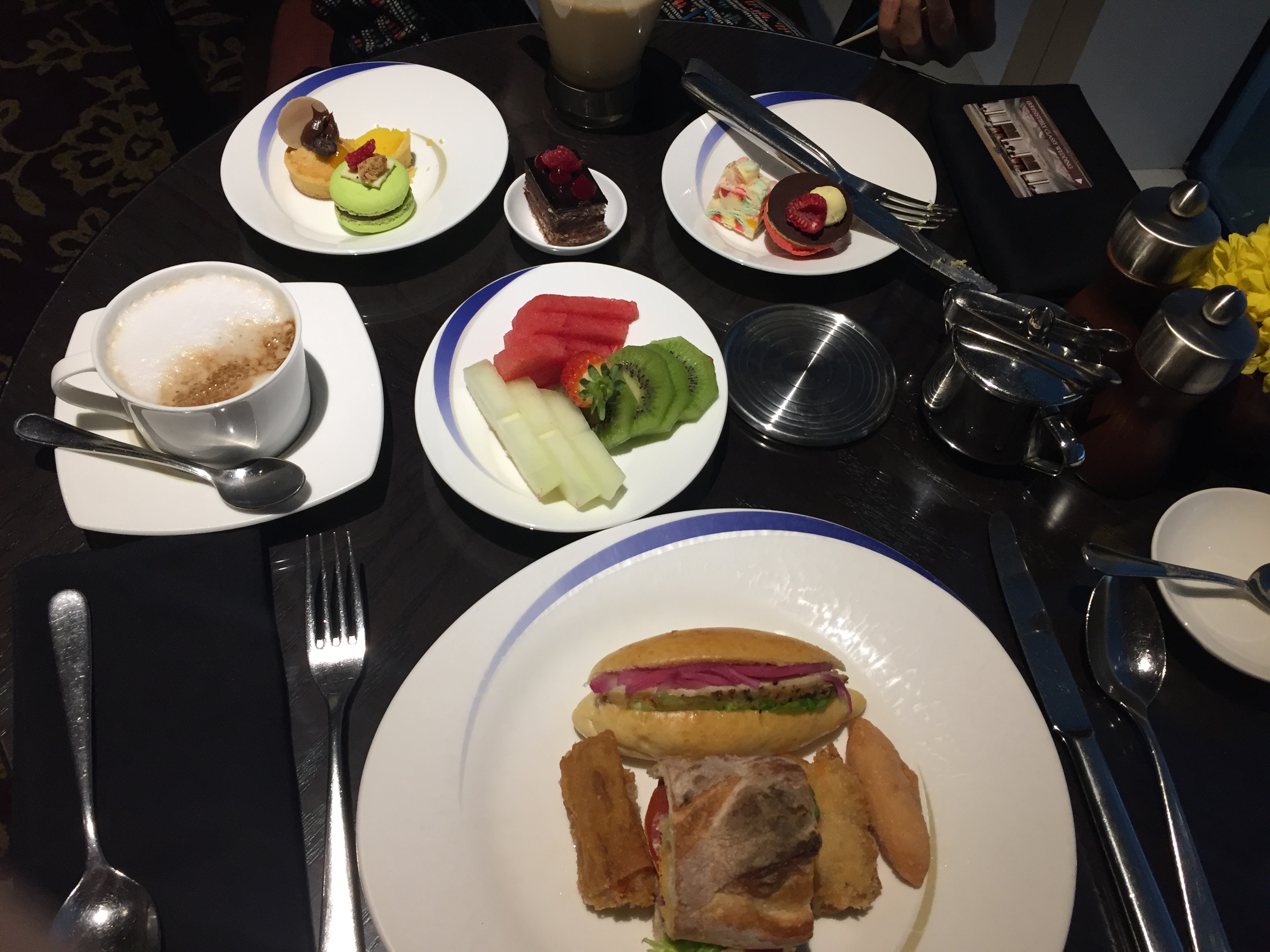 A plated afternoon tea — a slider with red onion, fresh fruit (watermelon, kiwi, melon, strawberry), a selection of sweets and macarons, finger sandwiches, and a cappuccino
