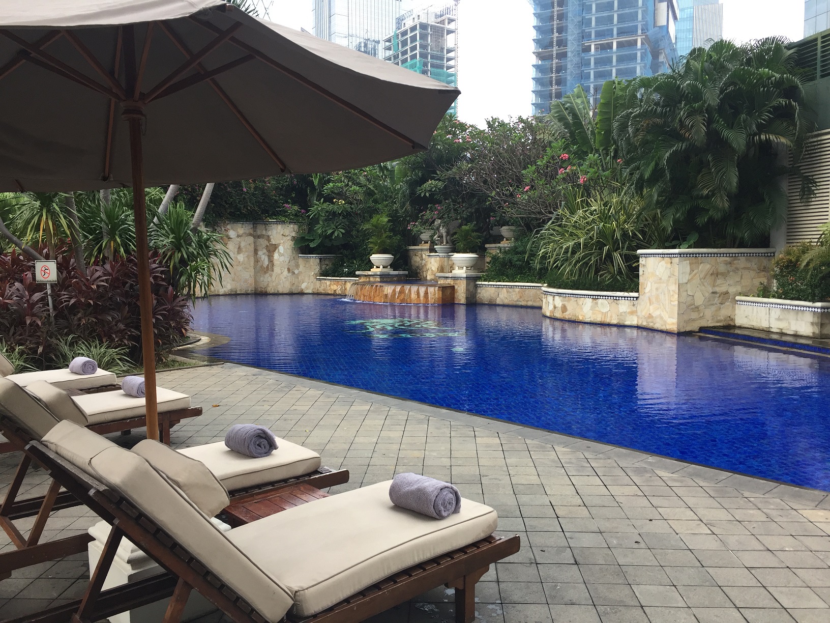 The hotel pool — deep blue water, lounge chairs with rolled towels, surrounded by tropical plantings and high-rises beyond