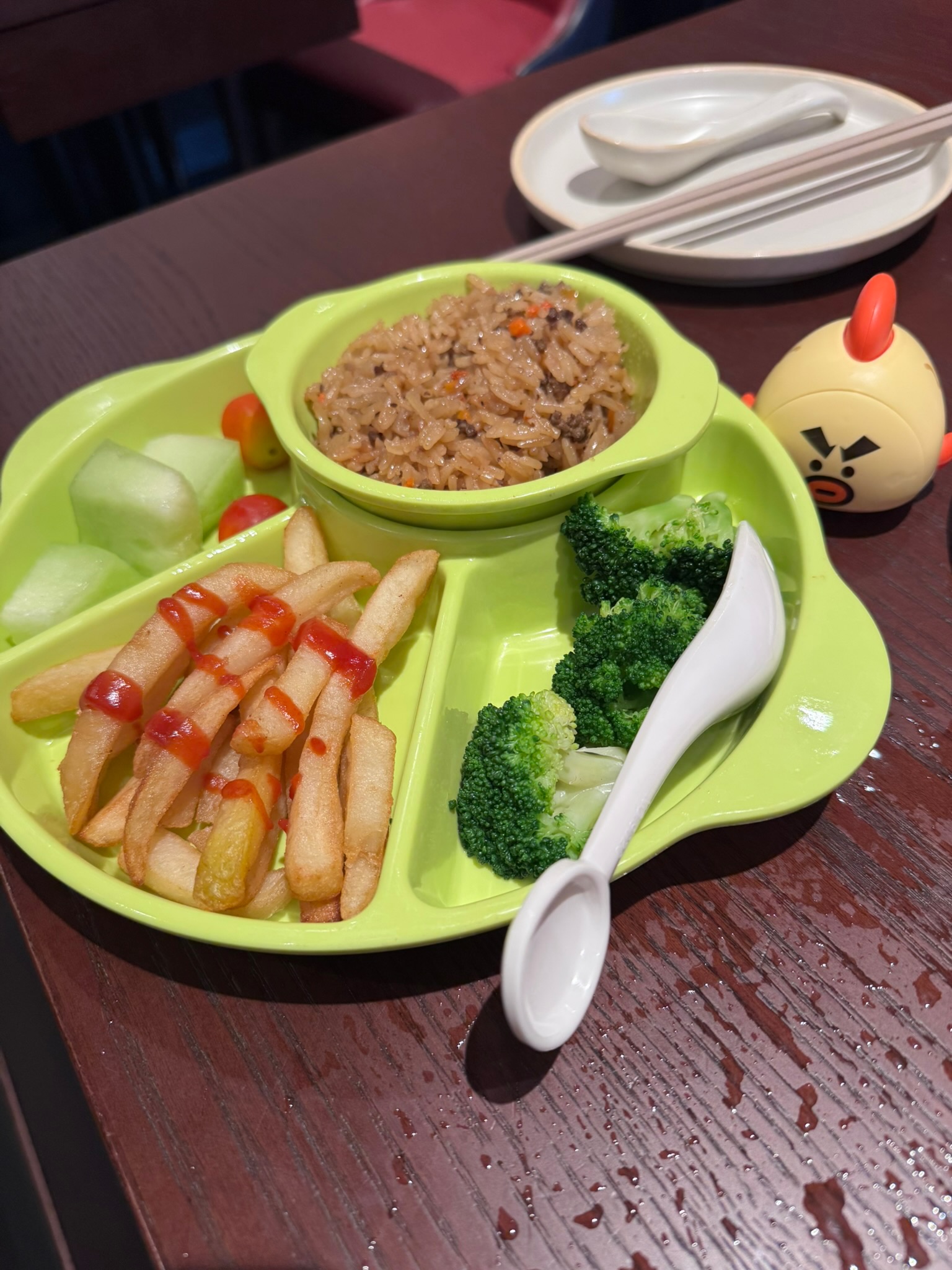 A bright green animal-shaped kid's plate with French fries (ketchup'd), broccoli, ground beef fried rice in a bowl, and melon cubes with cherry tomatoes — alongside a small chick-shaped toy