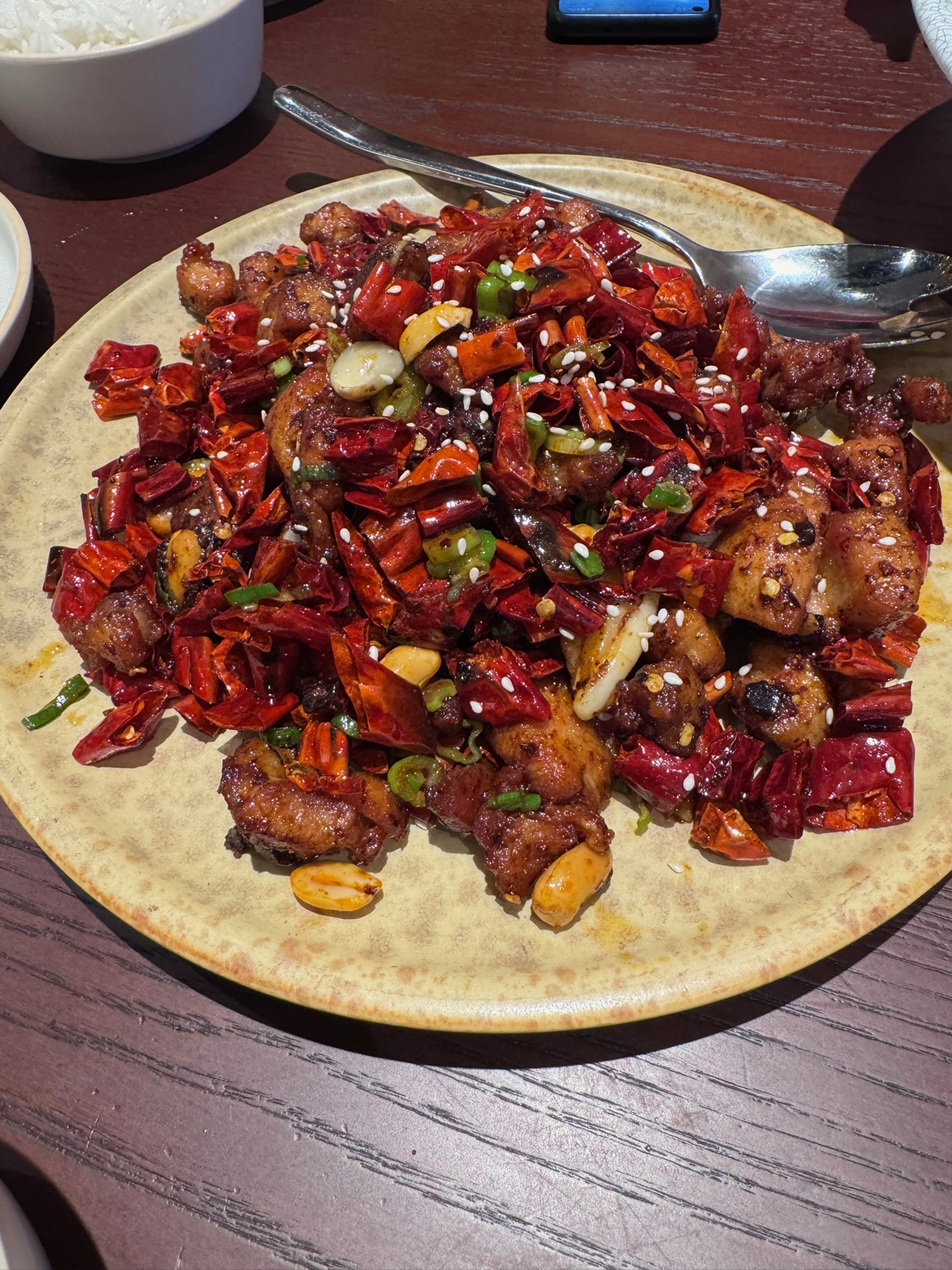 A plate of crispy fried chicken pieces almost completely buried under a mountain of dried red chilies, sesame seeds, scallions, and Sichuan peppercorns