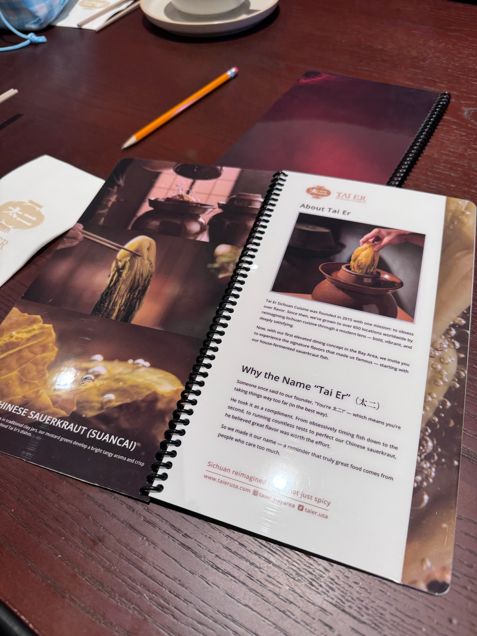 Menu pages spread across the table — "About Tai Er" with origin story, alongside a section about Chinese sauerkraut (suancai), with images of fermentation jars and noodles
