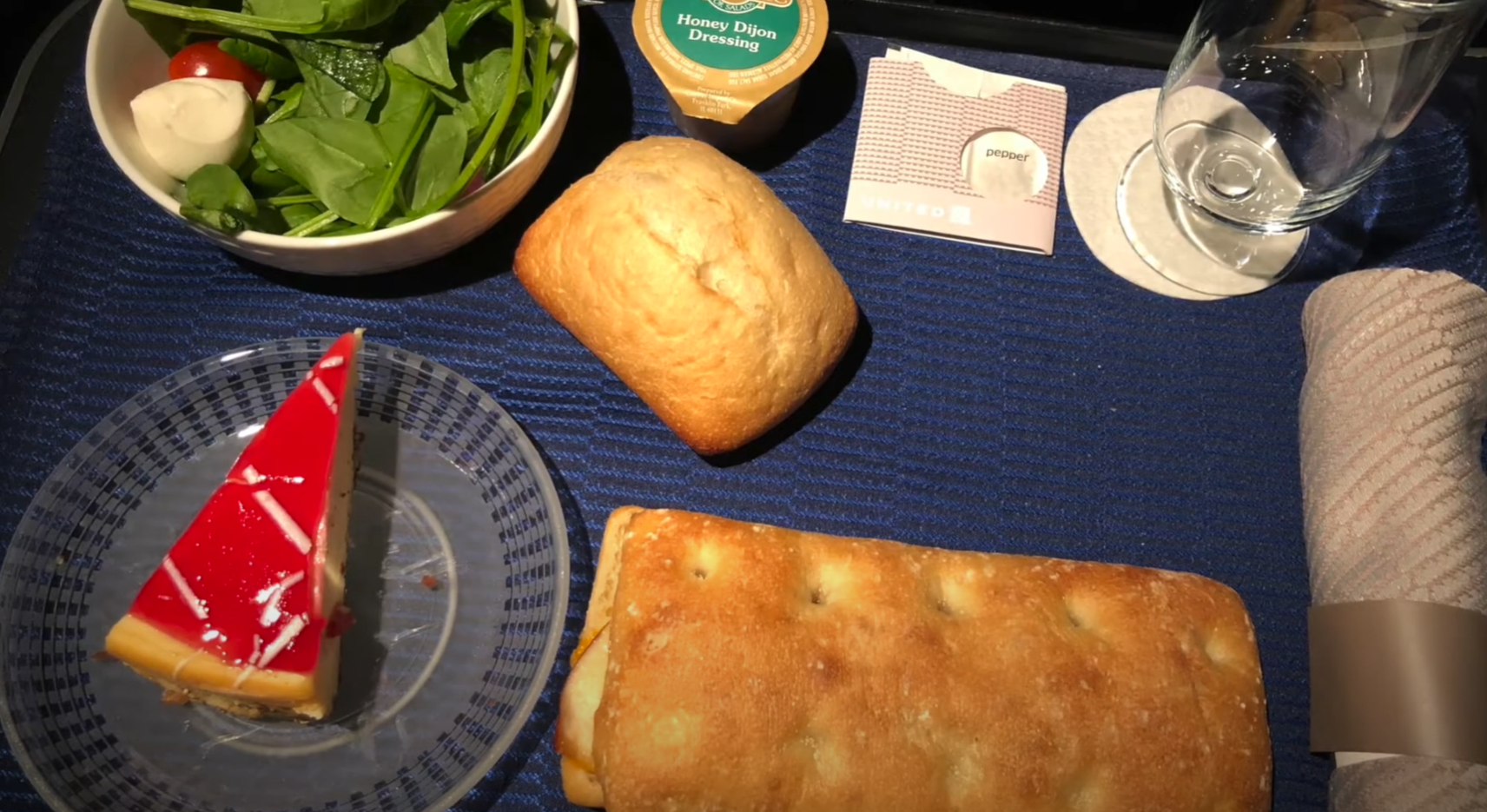 A domestic Business Class meal tray — a fresh salad with cherry tomatoes and mozzarella, a sealed packet of honey dijon dressing, a dinner roll, a slice of red-glazed cheesecake, a wrapped sandwich, a water glass, and a rolled napkin